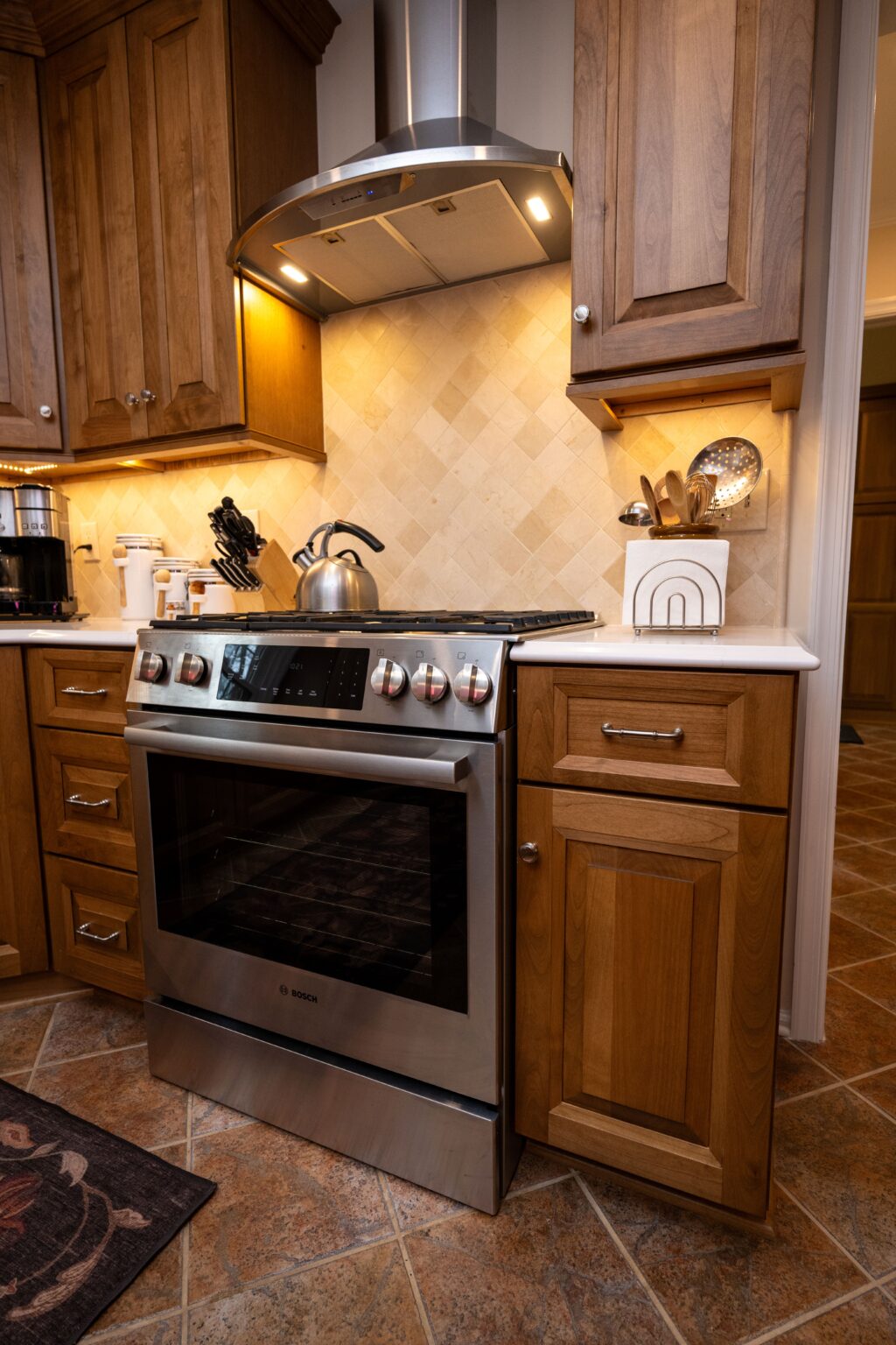 Stainless steel stove with kettle, surrounded by wooden cabinets and drawers in a tiled kitchen. Cooking utensils and appliances are on the countertop.
