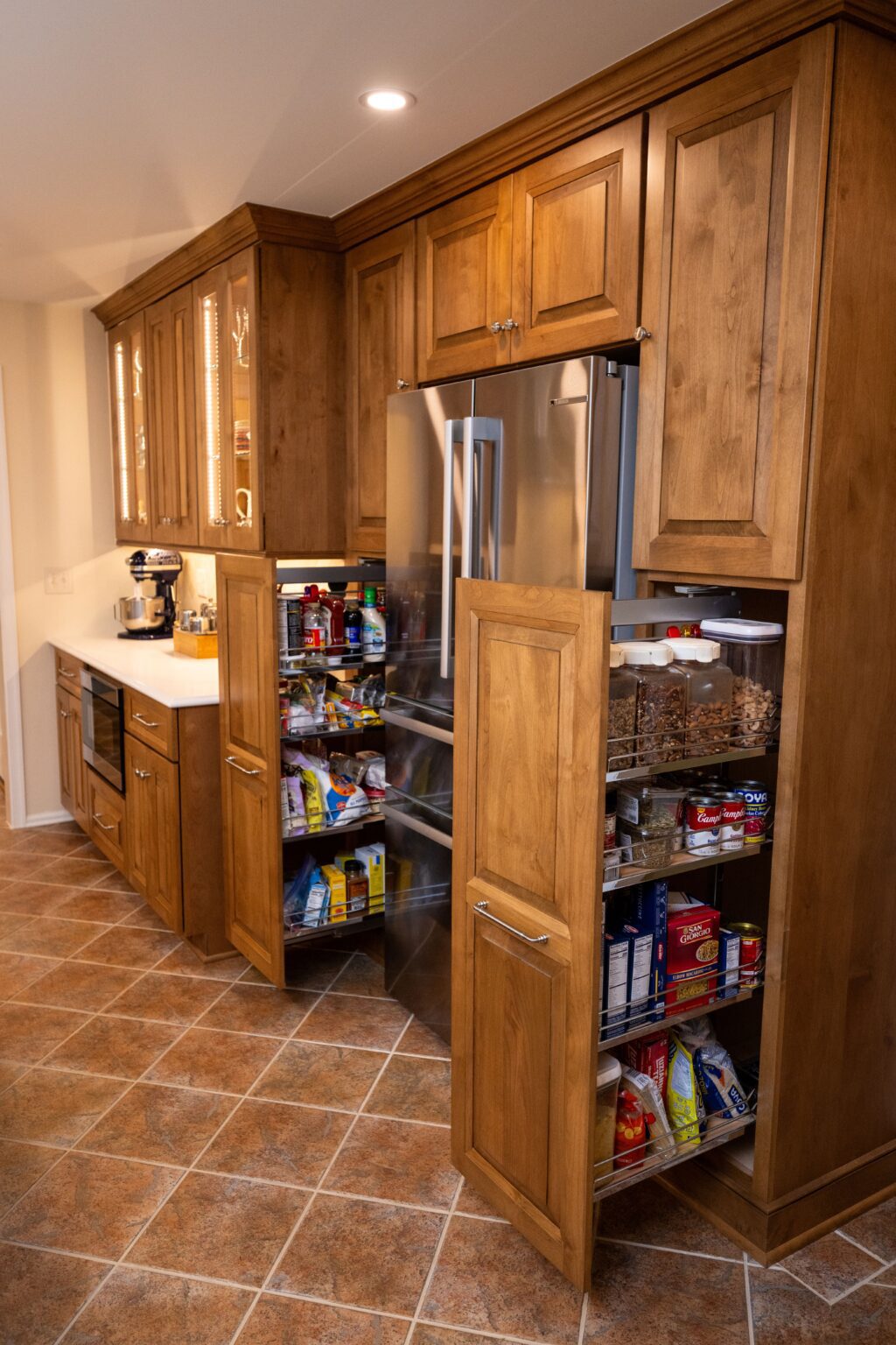 A kitchen with wood cabinets, a stainless steel refrigerator, and two pull-out pantry shelves stocked with various food items.