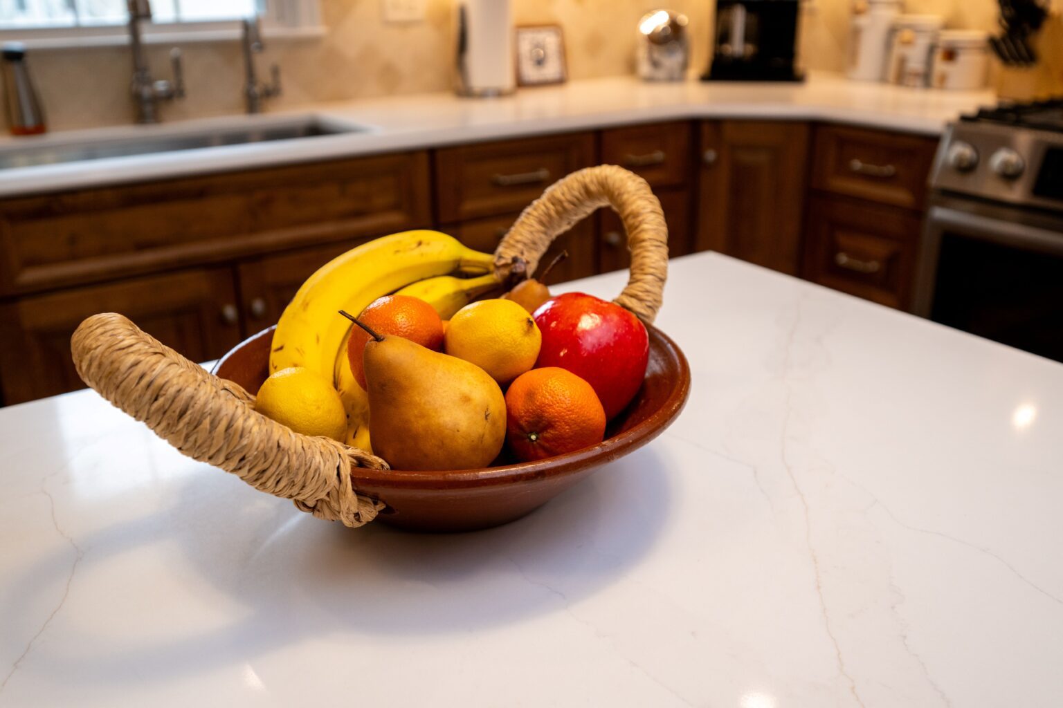 A brown bowl with bananas, a pear, apples, oranges, and a lemon sits on a white kitchen countertop.