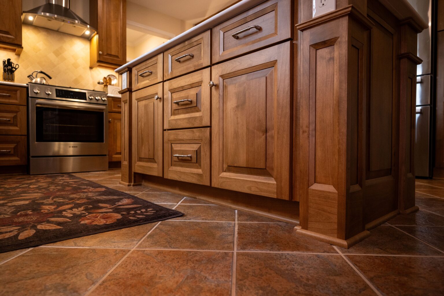 Close-up view of a kitchen island with wooden cabinets and drawers, tiled floor, and a stainless steel oven in the background.