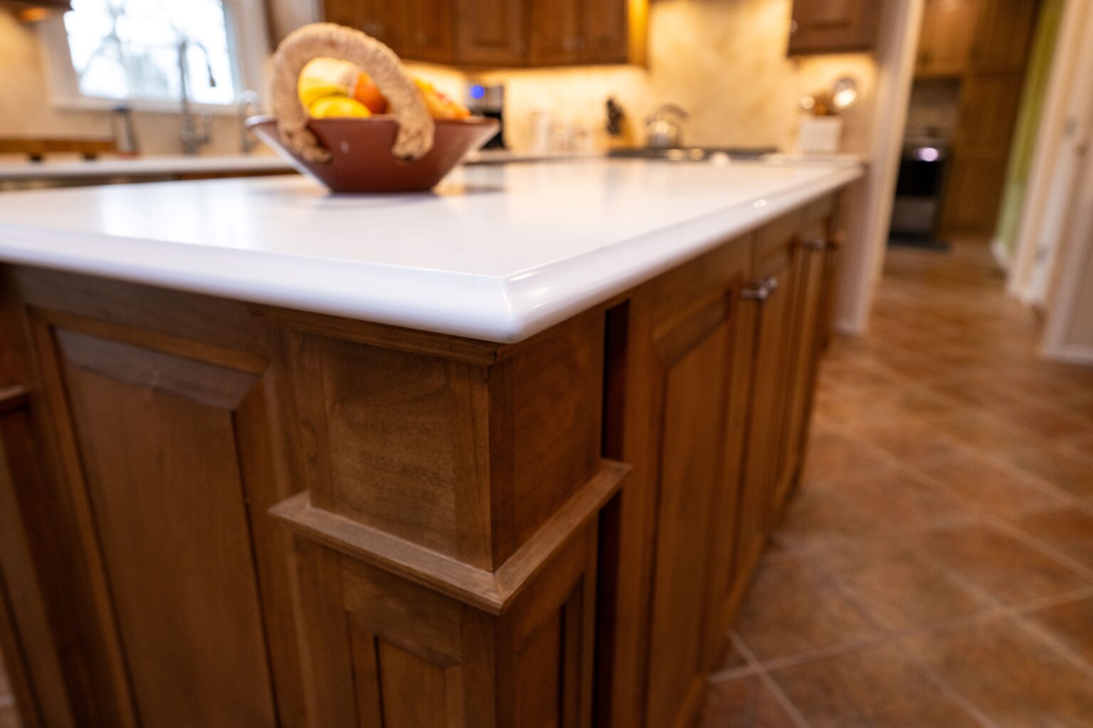 Close-up of a kitchen island with a white countertop and wooden cabinets, featuring a fruit bowl on top and tile flooring in the background.