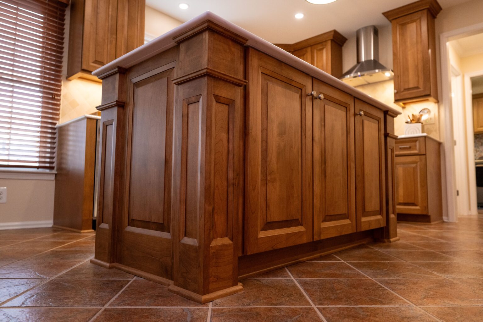 A wooden kitchen island with paneled cabinet doors stands on a tiled floor in a well-lit kitchen with wooden cabinetry.