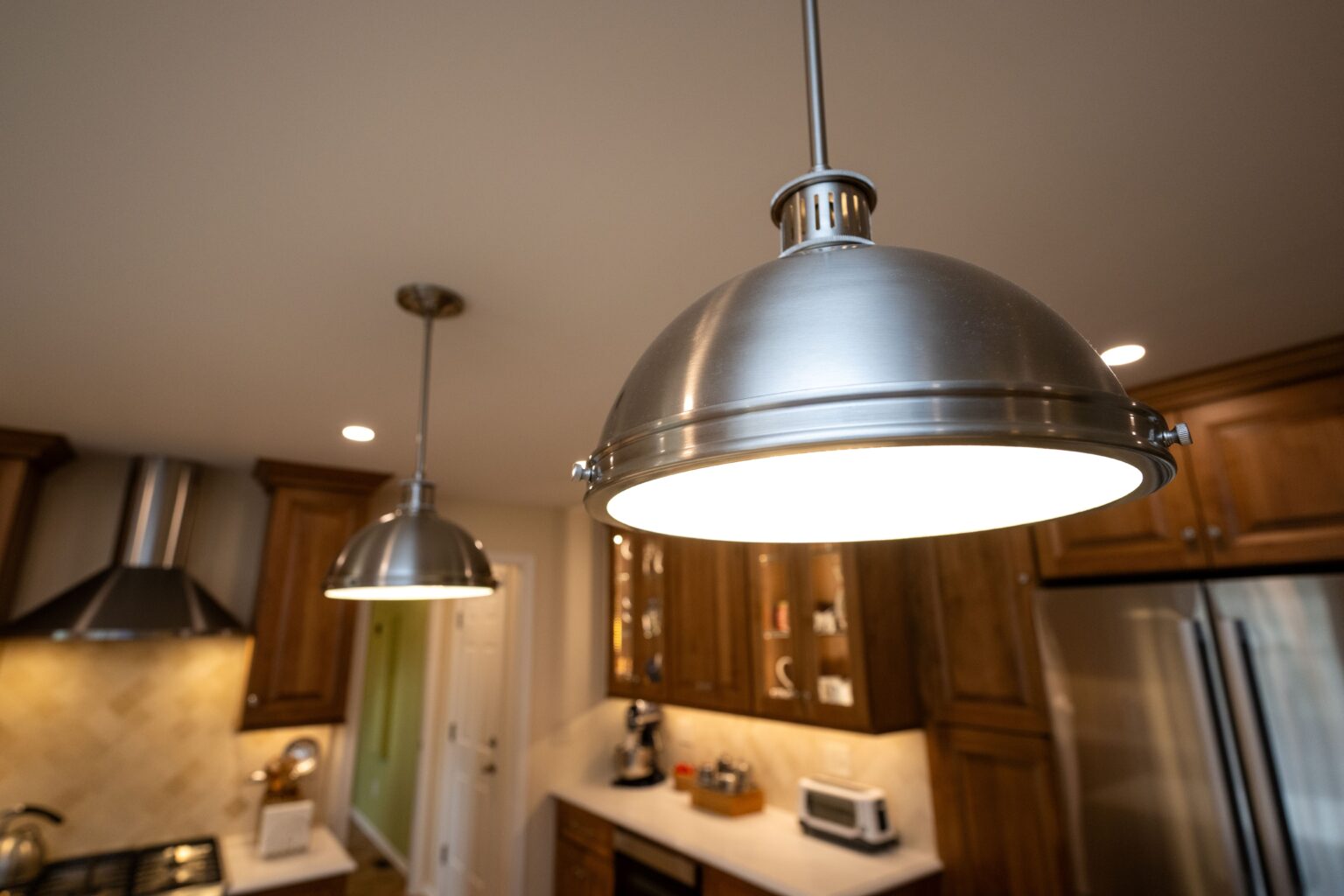 Close-up view of a modern pendant light fixture in a kitchen with wooden cabinets, stainless steel appliances, and a light-colored countertop.