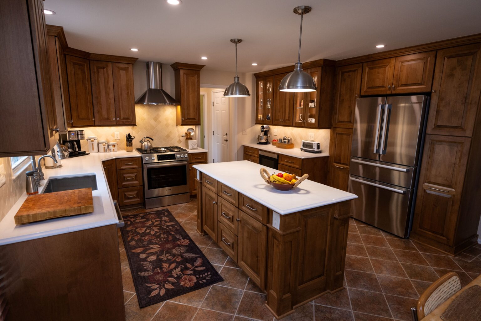 A modern kitchen with wooden cabinets, stainless steel appliances, a center island with a fruit basket, pendant lights, and tiled flooring.