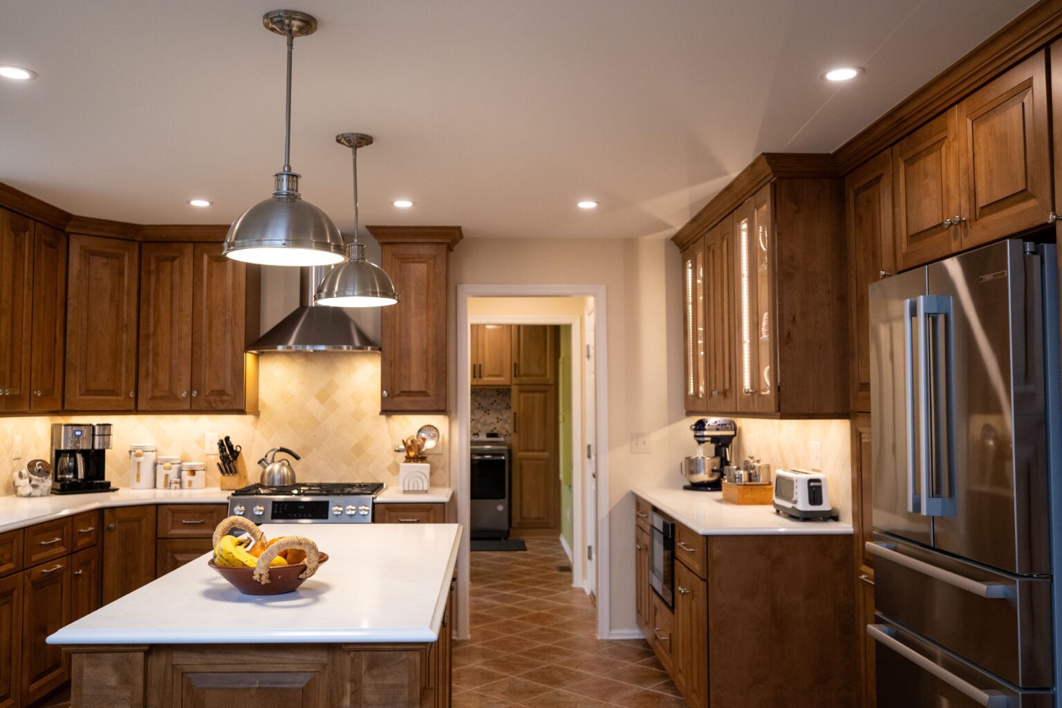 Modern kitchen with wooden cabinets, stainless steel appliances, white countertops, an island with a fruit bowl, and pendant lighting. A laundry room is visible through the doorway.