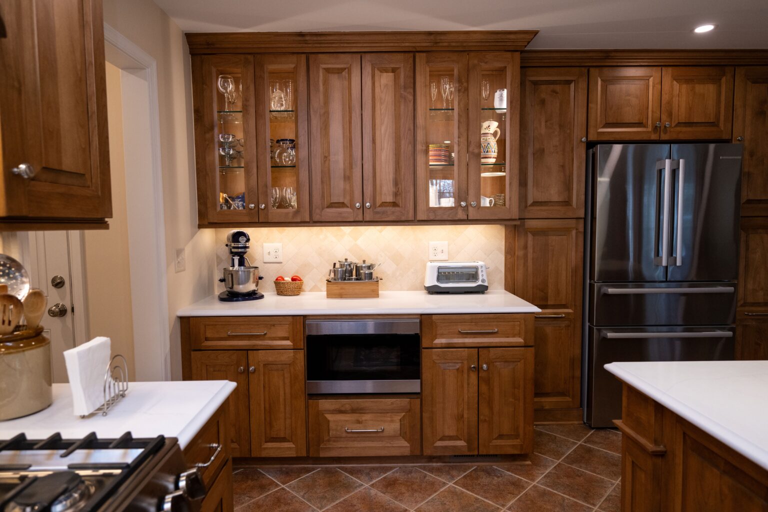 Kitchen with wooden cabinets, stainless steel refrigerator, oven, toaster, mixer, and countertop appliances. Warm lighting and tiled floor.