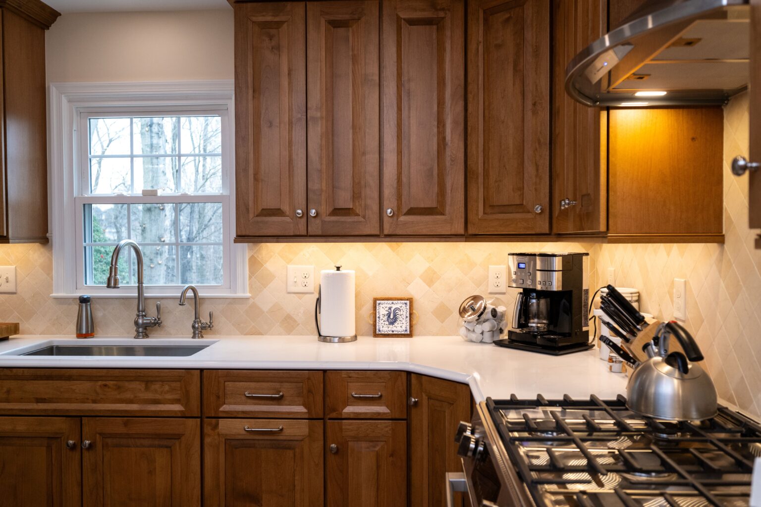 Modern kitchen with wooden cabinets, white countertops, a metal sink, a coffee maker, a paper towel holder, a knife block, and a stove with a kettle under warm lighting.