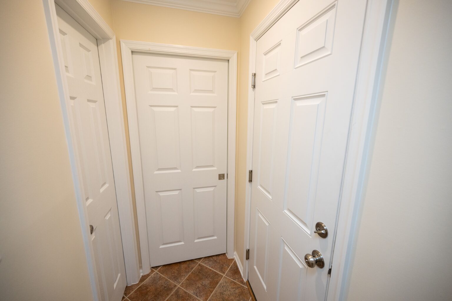 A small hallway with three closed white doors and beige walls, featuring brown and tan tiled flooring.