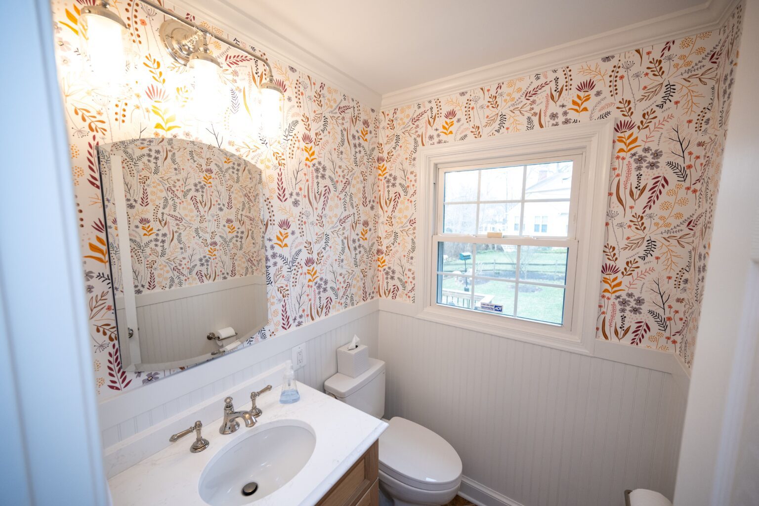 Small bathroom with floral wallpaper, white sink and countertop, wall-mounted mirror, toilet, and window with outdoor view. Bright lighting above the mirror.