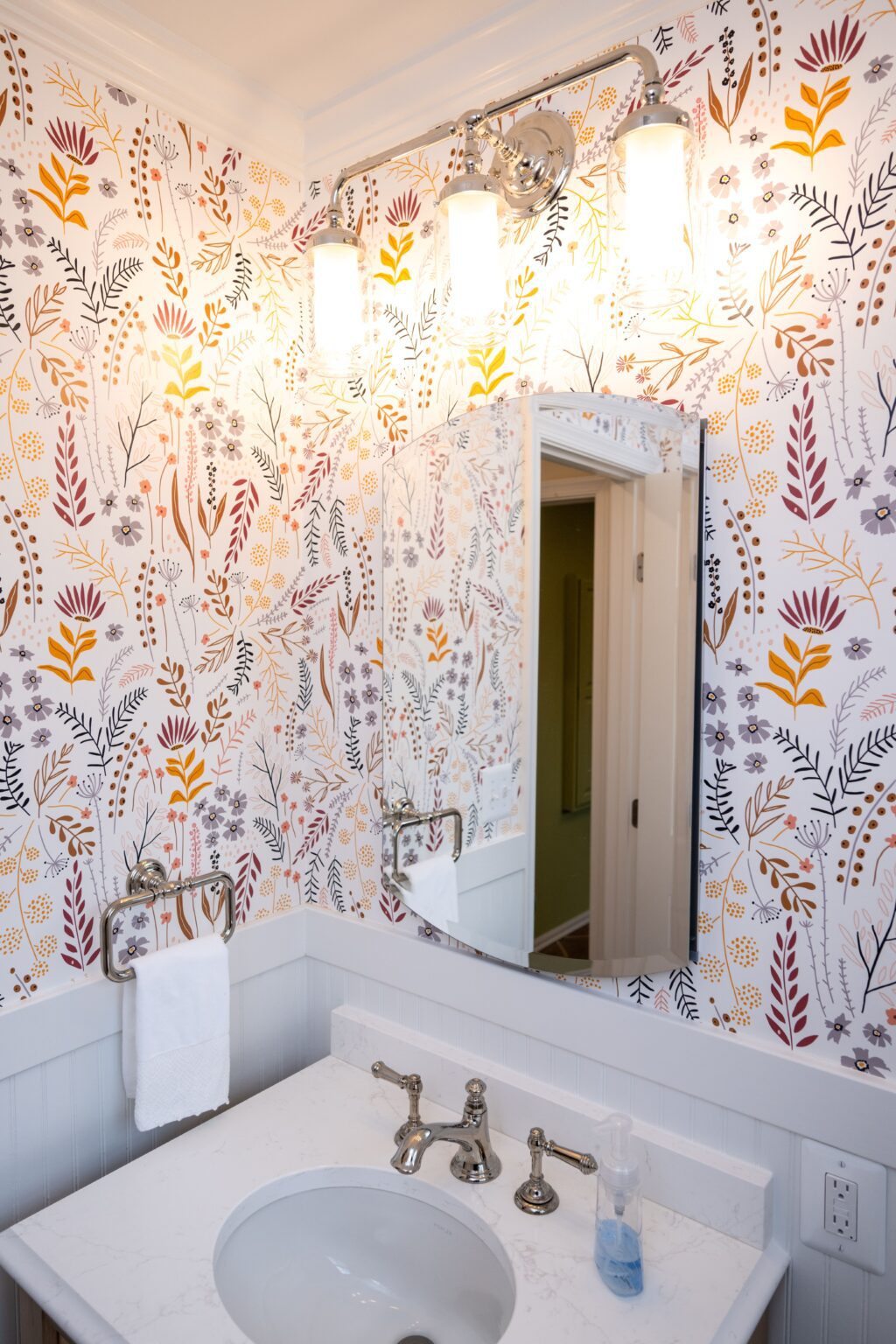 Bathroom sink and mirror with a floral patterned wallpaper, towel on a rack, soap dispenser on the counter, and three lit vanity lights above the mirror.