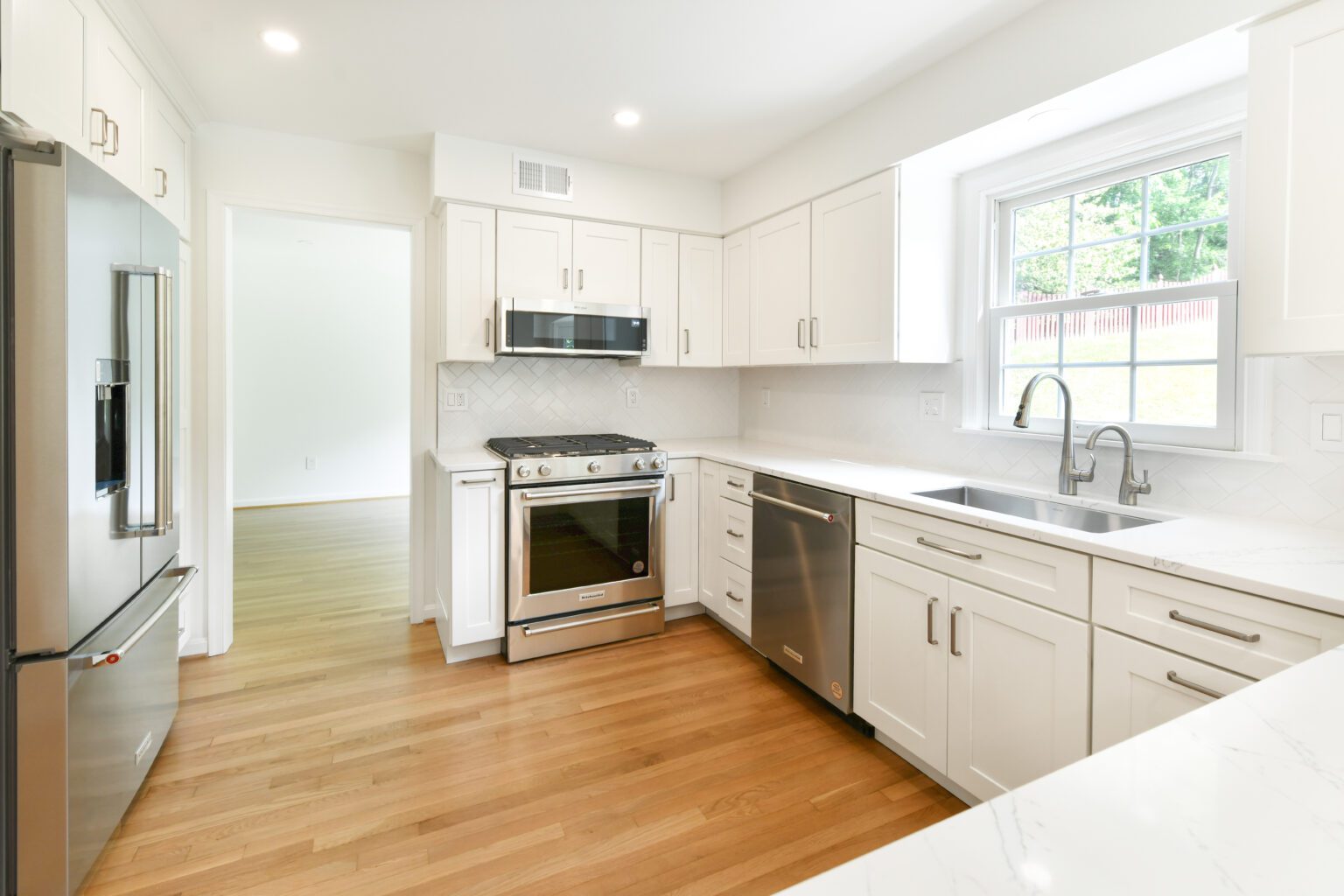 Modern kitchen with white cabinets, stainless steel appliances, a large window above the sink, and light wood flooring.