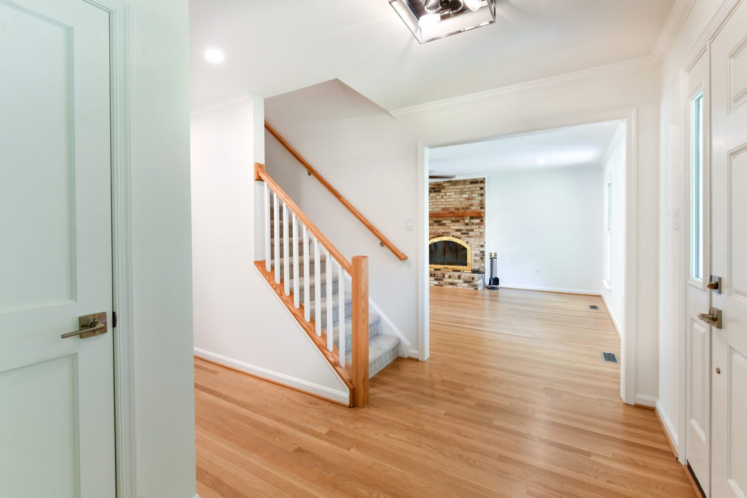 Entryway with hardwood floors, a staircase with white spindles and wooden handrail, and a view into a living room with a brick fireplace.