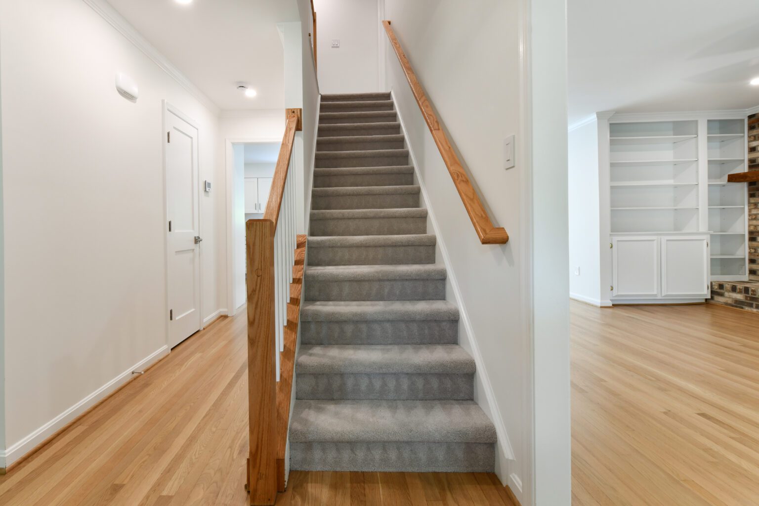 Carpeted staircase with wooden handrails leading upward between white walls, adjacent to a hallway with light hardwood floors and built-in shelves visible in the next room.