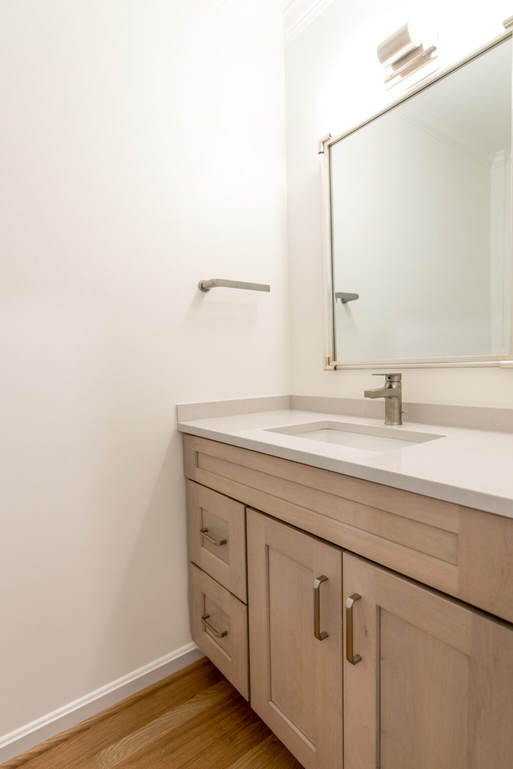 Modern bathroom with a light wood vanity, white countertop, sink, wall-mounted faucet, large mirror, towel bar, and hardwood floor.