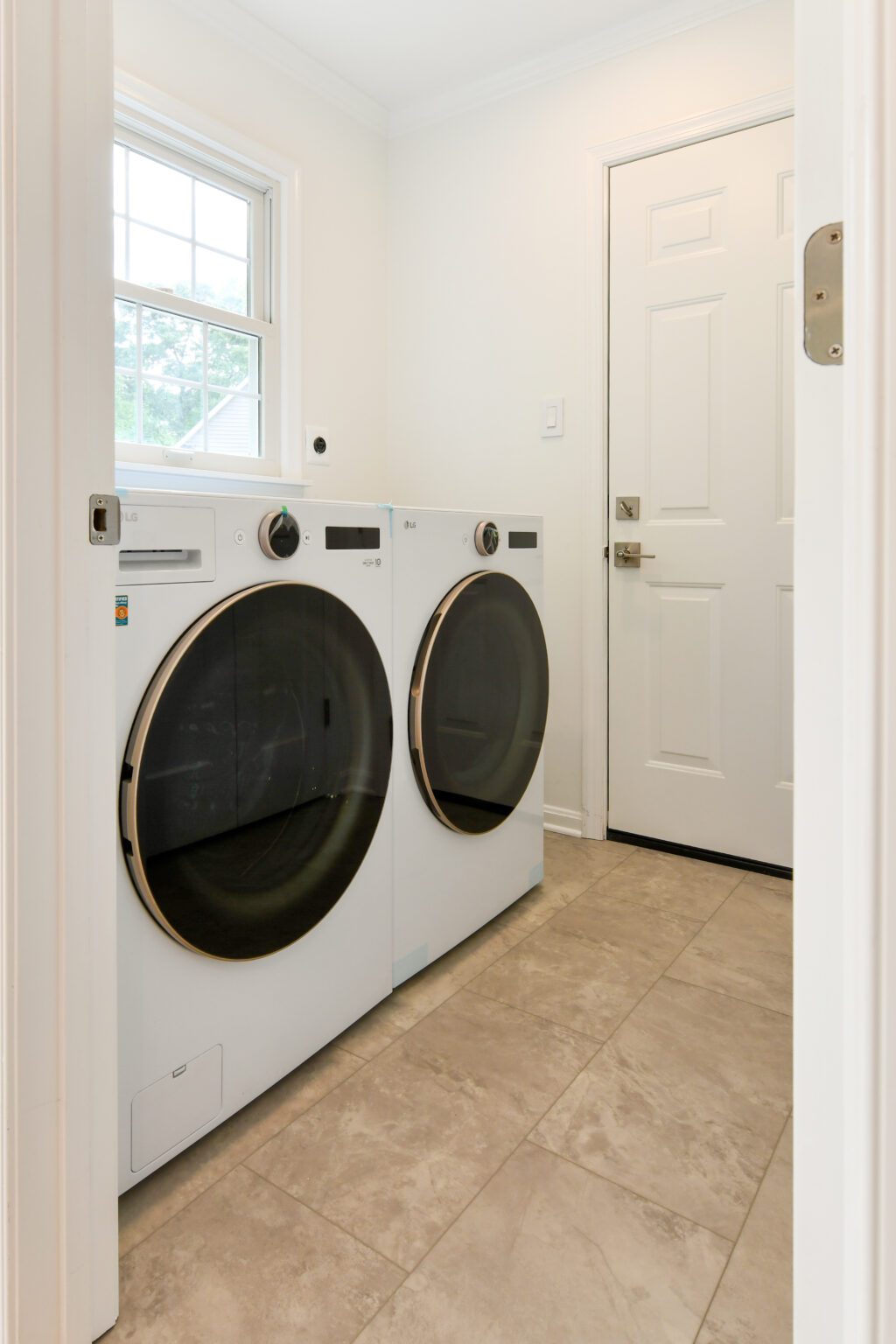 A laundry room with a front-loading washer and dryer side by side on a tiled floor, with a window and a door in the background.
