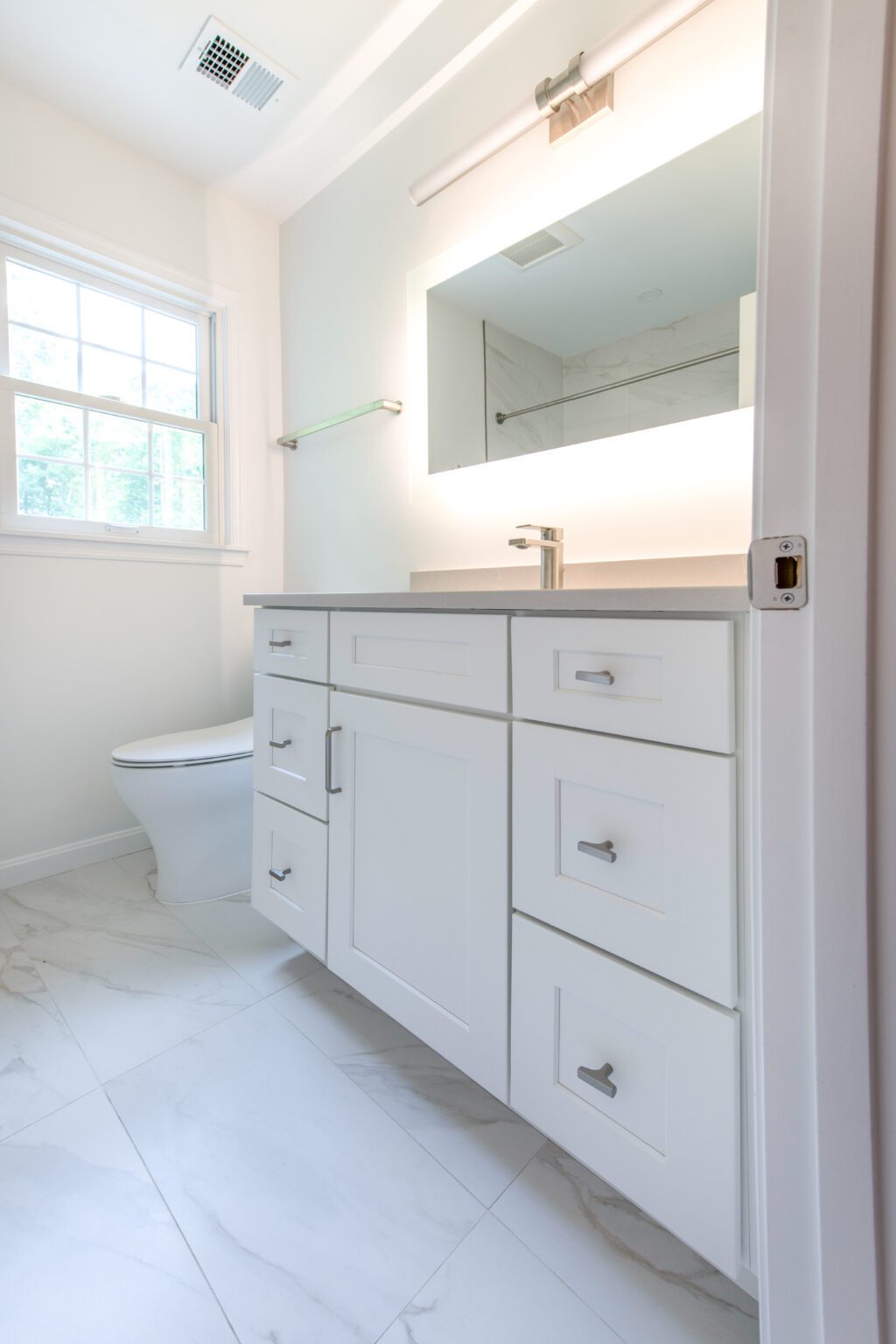 Modern bathroom with white cabinetry, a single-handle faucet, under-cabinet lighting, a window, and a toilet on white marble tile flooring.