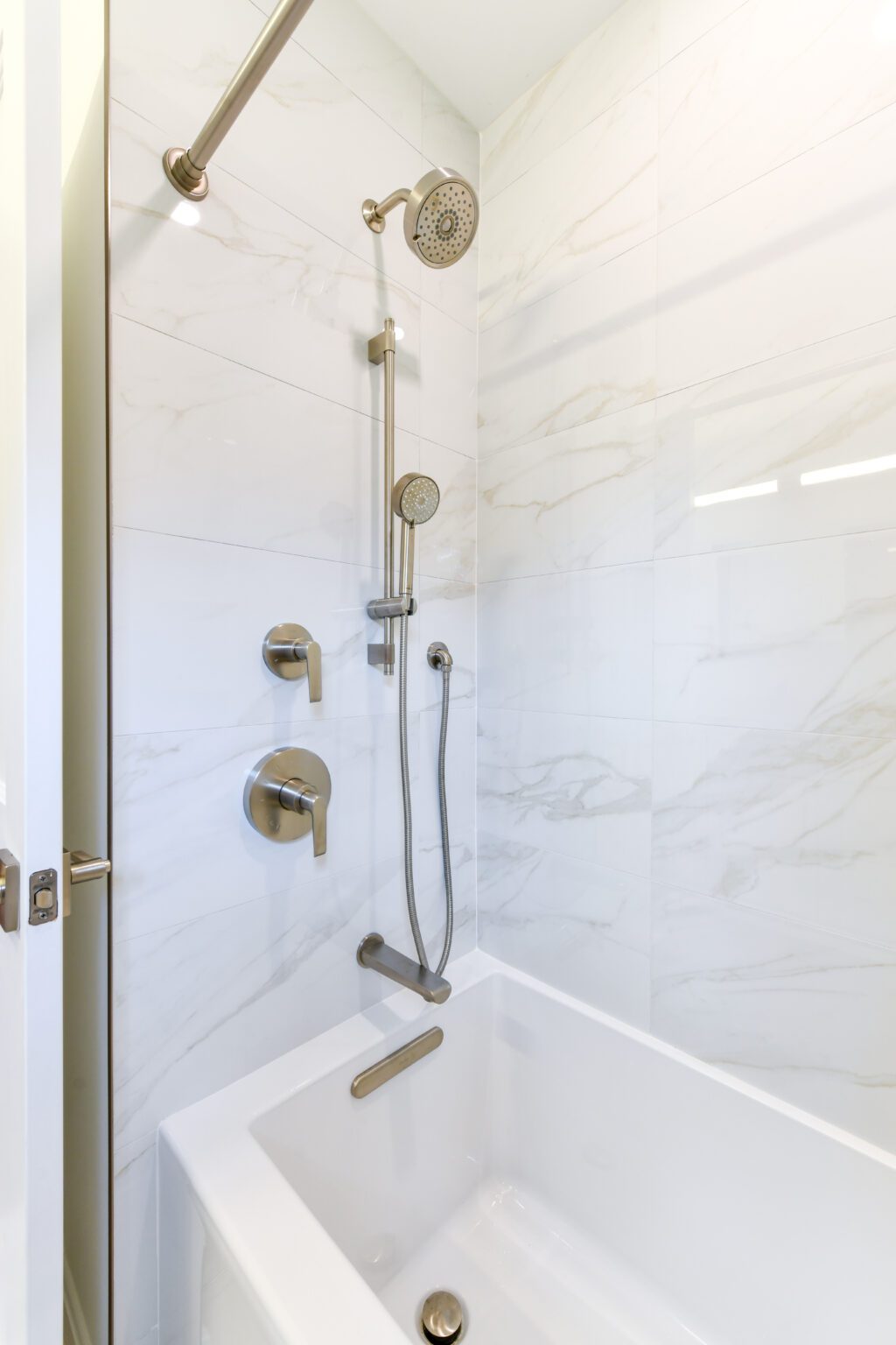 Modern white bathtub shower with marble-patterned tile wall, featuring a rain showerhead, handheld shower, and metal fixtures.