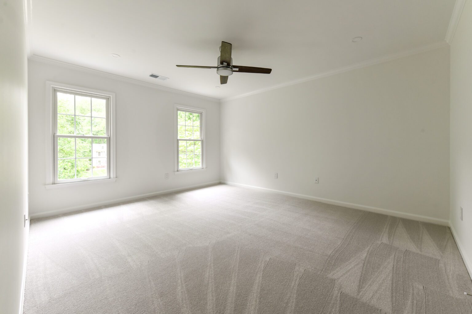 Empty room with light-colored carpet, white walls, two large windows, and a ceiling fan with a light fixture.