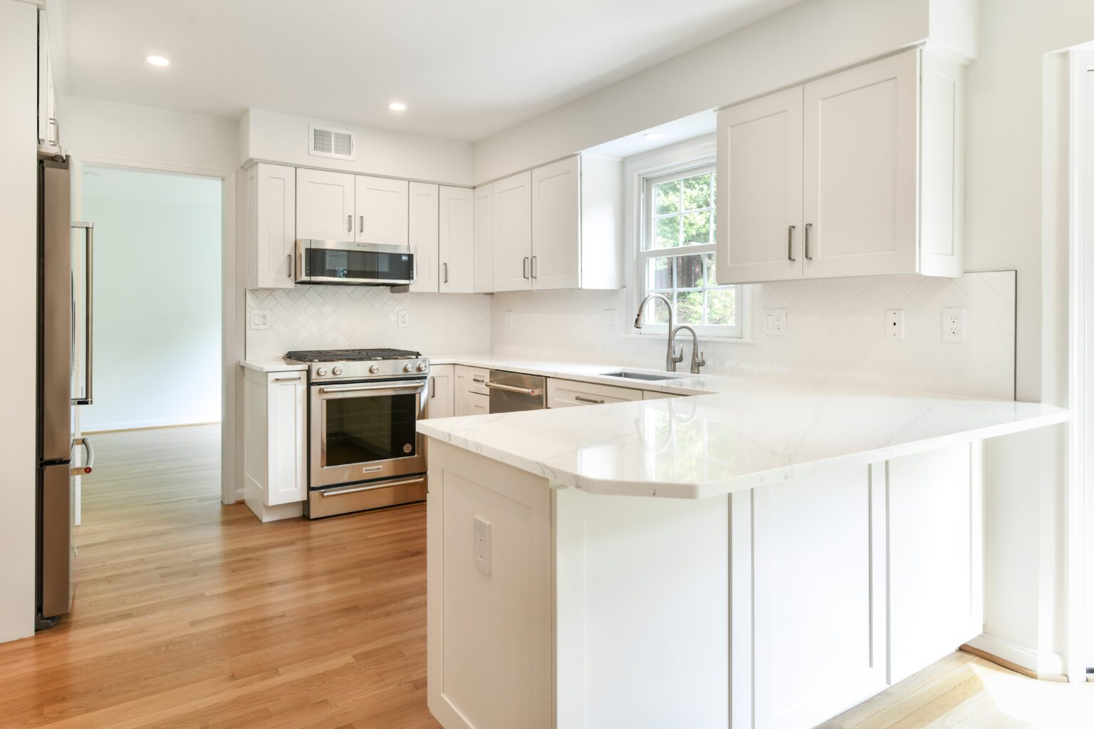 Modern kitchen with white cabinets, stainless steel appliances, a large window above the sink, and light wood flooring.