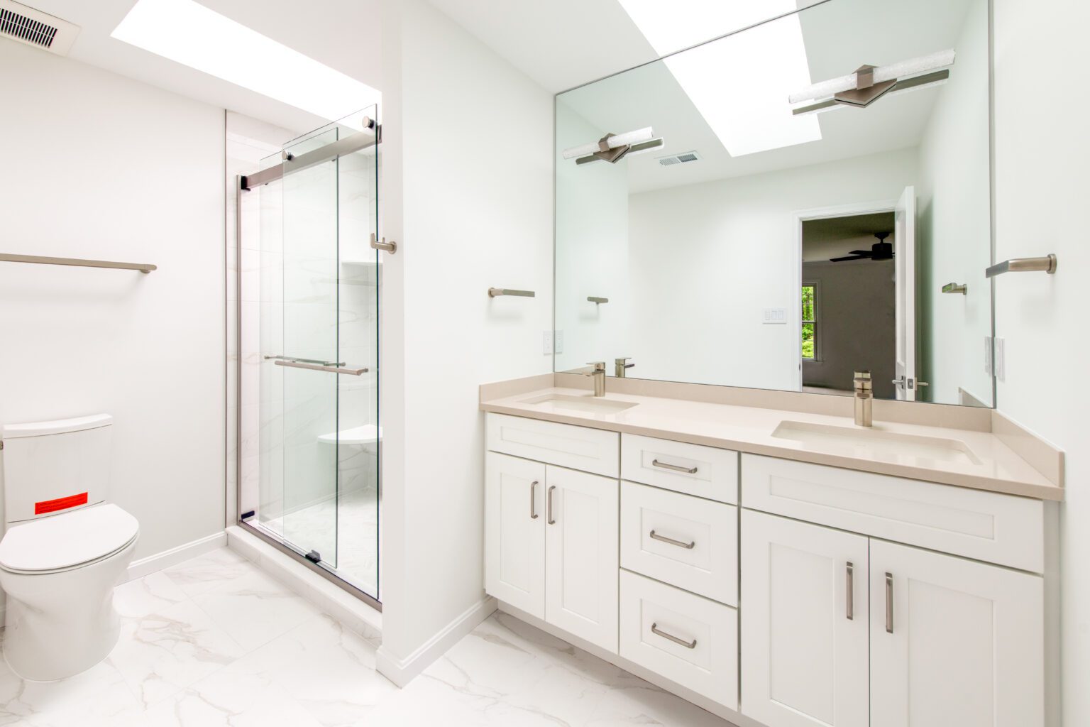 Modern bathroom with white cabinets, double sink vanity, large mirror, glass shower enclosure, toilet, and marble tile floor under skylights.