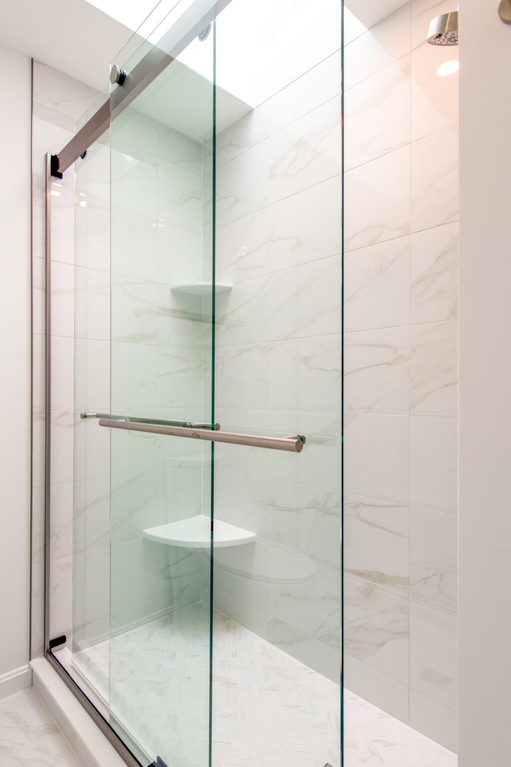 Modern shower with sliding glass doors, chrome handle, white marble-patterned tile walls, and two corner shelves.