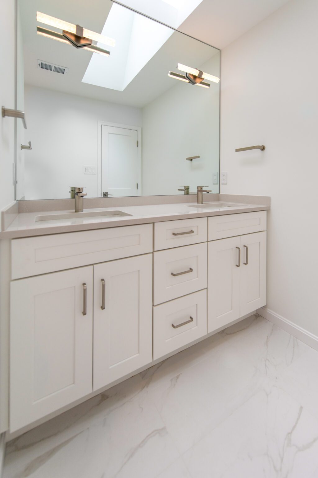 White bathroom vanity with dual sinks, large mirror, modern fixtures, and marble-patterned floor tiles under a skylight.