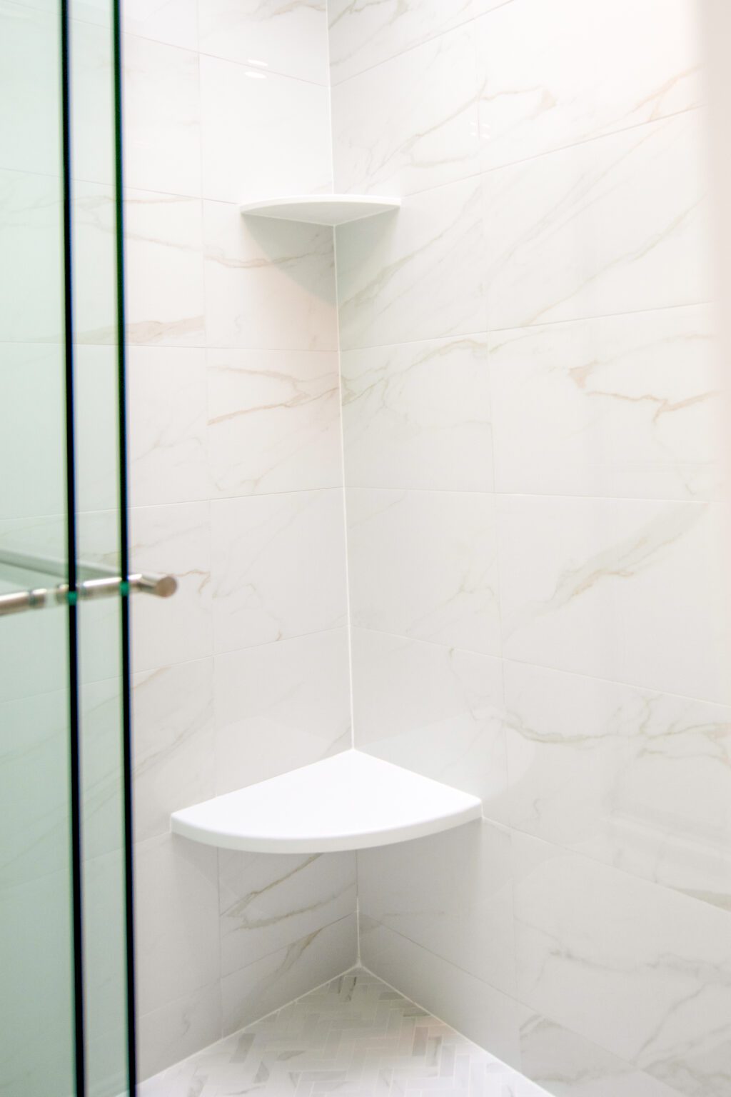 A shower corner with white marble-patterned tiles, a glass door, and two white triangular shelves mounted in the corner.
