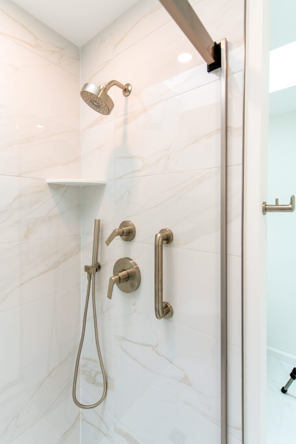 Modern shower with white marble tile, metal showerhead, handheld sprayer, two control knobs, grab bar, and a corner shelf.