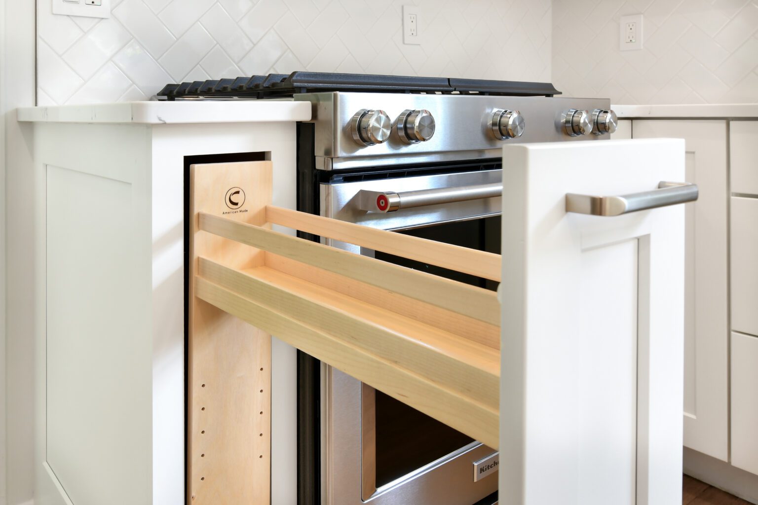 A close-up of a modern kitchen with a pull-out wooden spice rack drawer next to a stainless steel stove and white cabinetry.