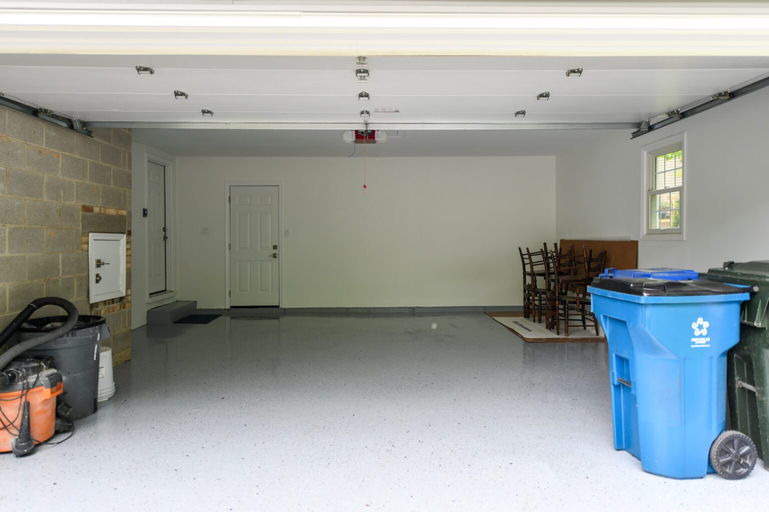 A clean, empty garage with a few stacked chairs and a table in the corner, trash and recycling bins, cleaning supplies, and a partially open garage door.