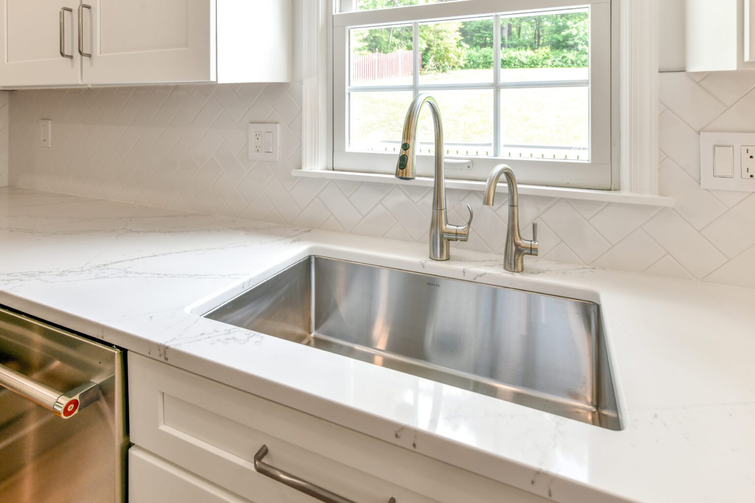A modern kitchen sink with a stainless steel basin and two faucets, set in a white countertop beneath a window with a view of grass and trees outside.