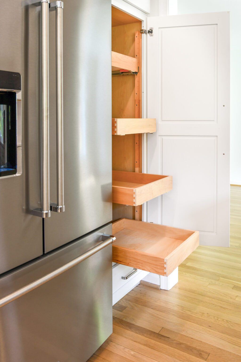 A kitchen cabinet with three wooden pull-out shelves is open next to a stainless steel refrigerator on a light wood floor.