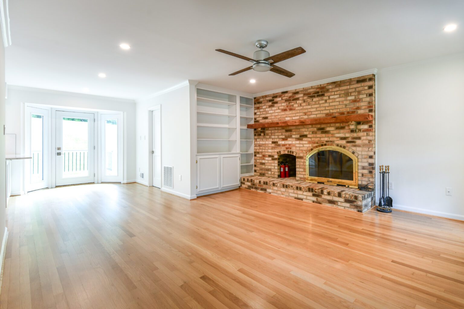 Spacious living room with light wood floors, brick fireplace, built-in shelves, ceiling fan, and double glass doors leading outside.