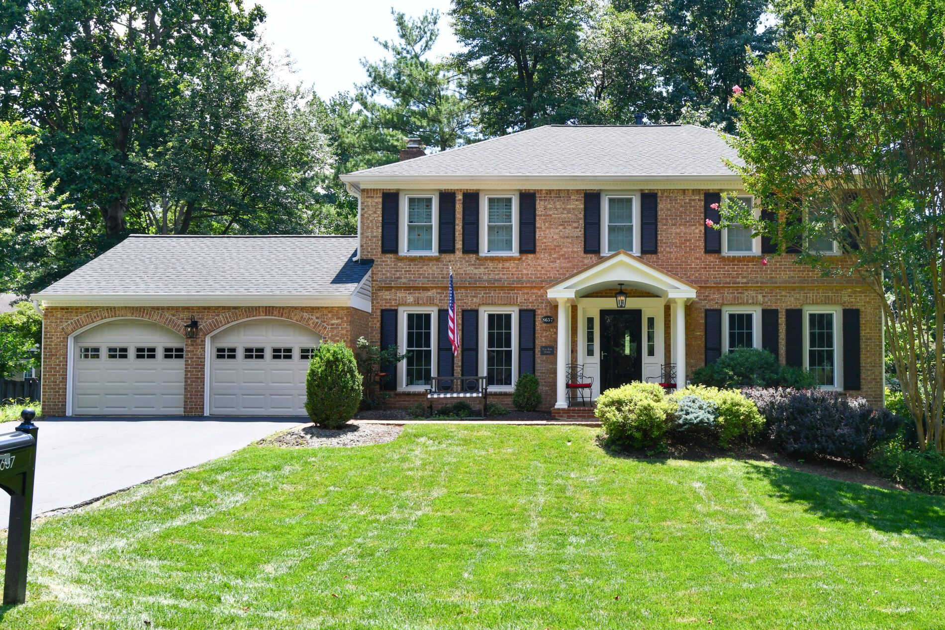 Two-story brick house with black shutters, a double garage, a well-maintained lawn, and landscaped bushes, set against a backdrop of tall trees.