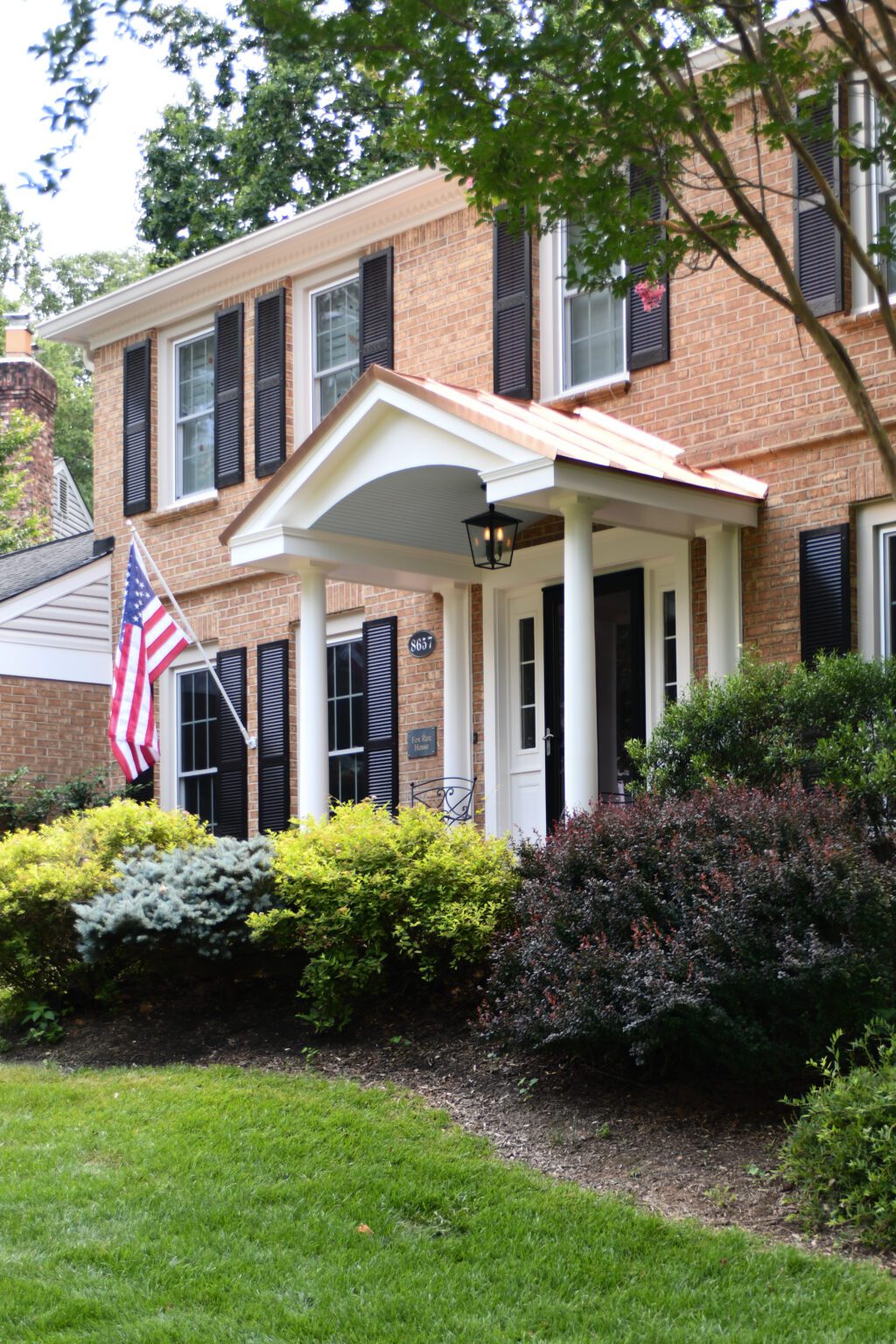 Vienna Metal Roof Portico (10) Two-story brick house with black shutters, a covered front porch, an American flag, and landscaped front yard with green bushes and grass.