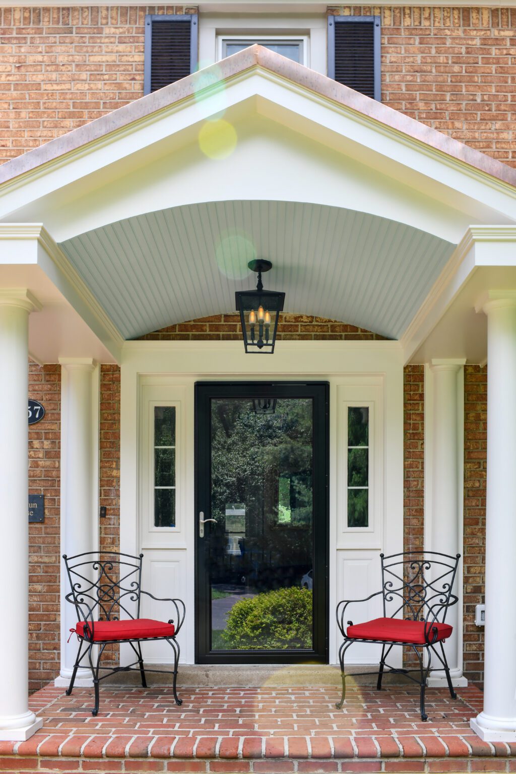 Vienna Metal Roof Portico (14) A front porch with two black chairs with red cushions, brick steps, white columns, and a black framed glass door under a peaked roof with a hanging lantern.