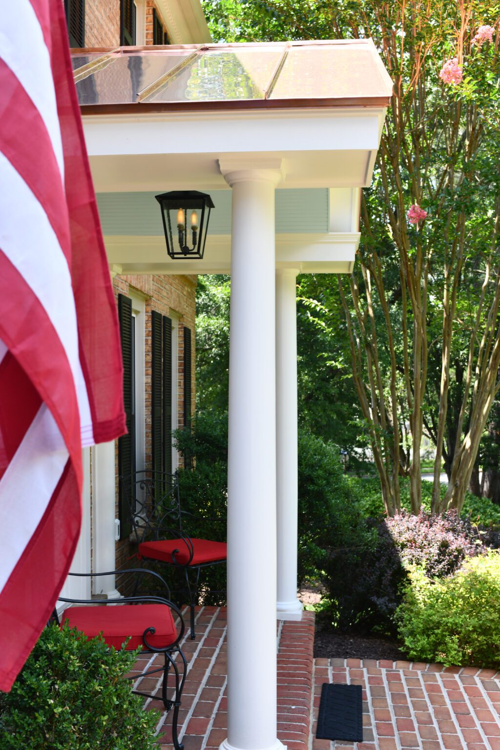 Vienna Metal Roof Portico (2) A brick front porch with white columns, red-cushioned chairs, a hanging lantern, a flowering tree, and an American flag in the foreground.
