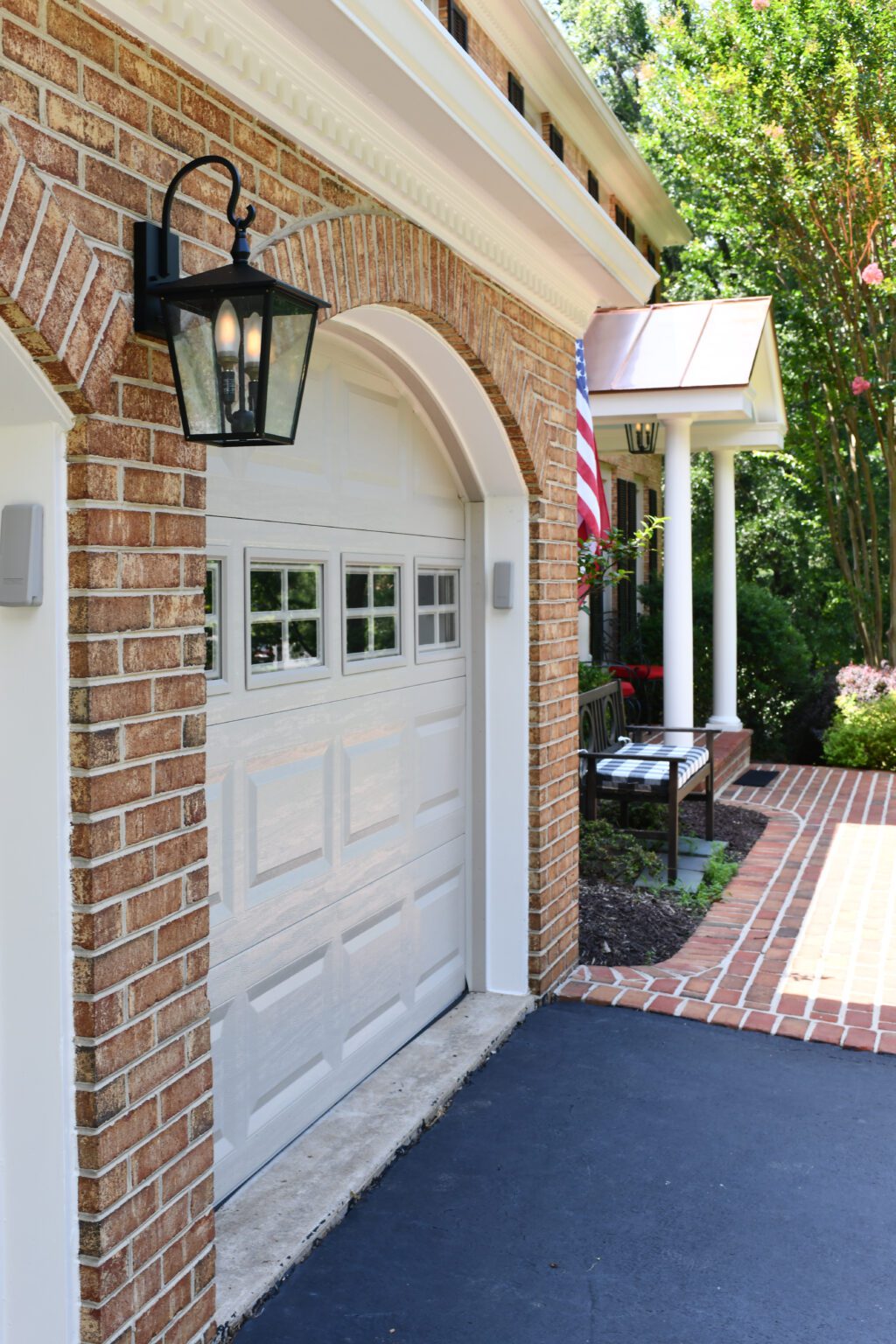 Vienna Metal Roof Portico (4) A white garage door with small windows is set in a brick wall, next to a black lantern and an American flag, with a walkway leading to a porch.