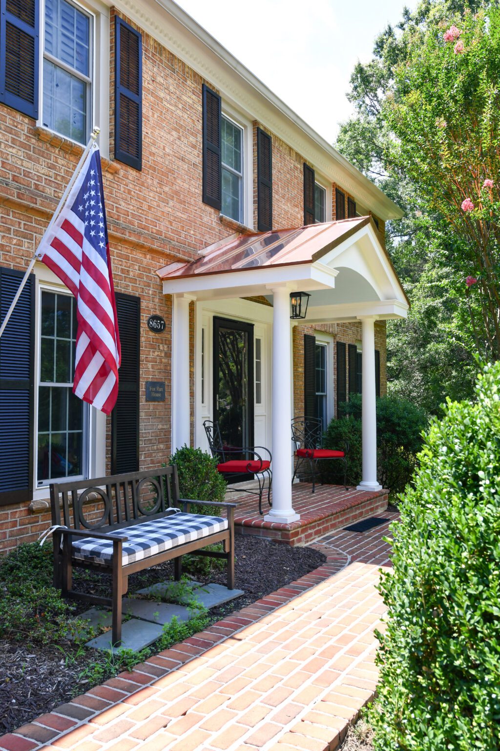 Vienna Metal Roof Portico (5) A brick house with black shutters, a front porch with white columns, two chairs with red cushions, a bench with striped cushion, and an American flag displayed.