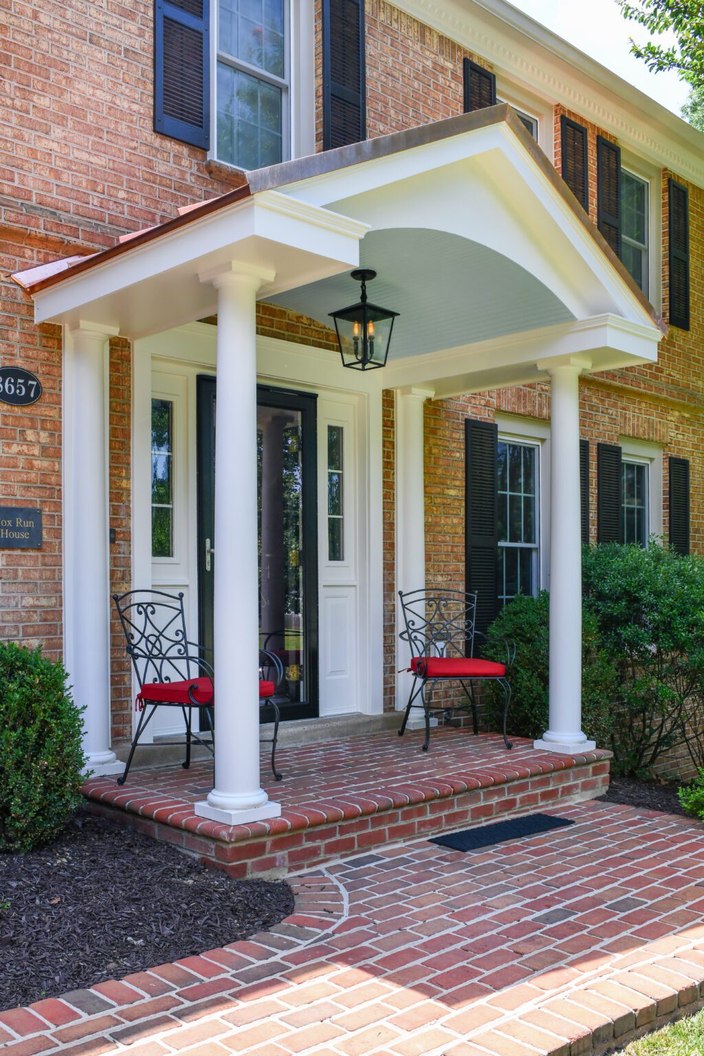 Vienna Metal Roof Portico (6) Brick house entryway with two white columns, a covered porch, two black chairs with red cushions, brick steps, and black shutters on the windows.