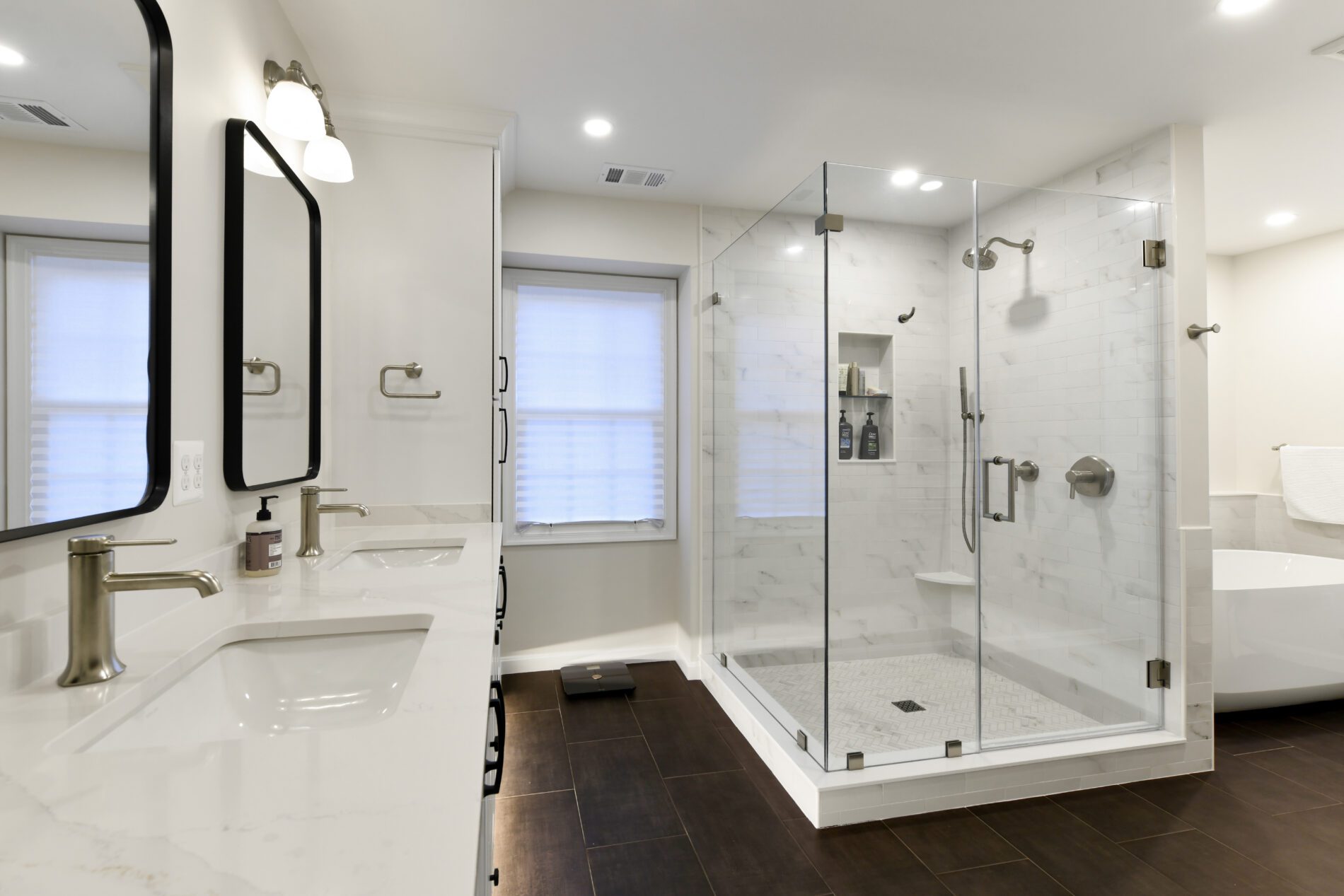 Modern bathroom with double sinks, brass fixtures, large glass-enclosed shower, white countertops, freestanding tub, and dark wood floor.
