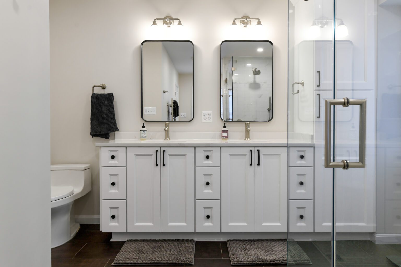 Vienna Primary En-suite and Laundry (11) Modern bathroom with double vanity, two mirrors, overhead lights, white cabinets, a toilet on the left, and a glass shower door on the right. Two gray floor mats are in front of the vanity.