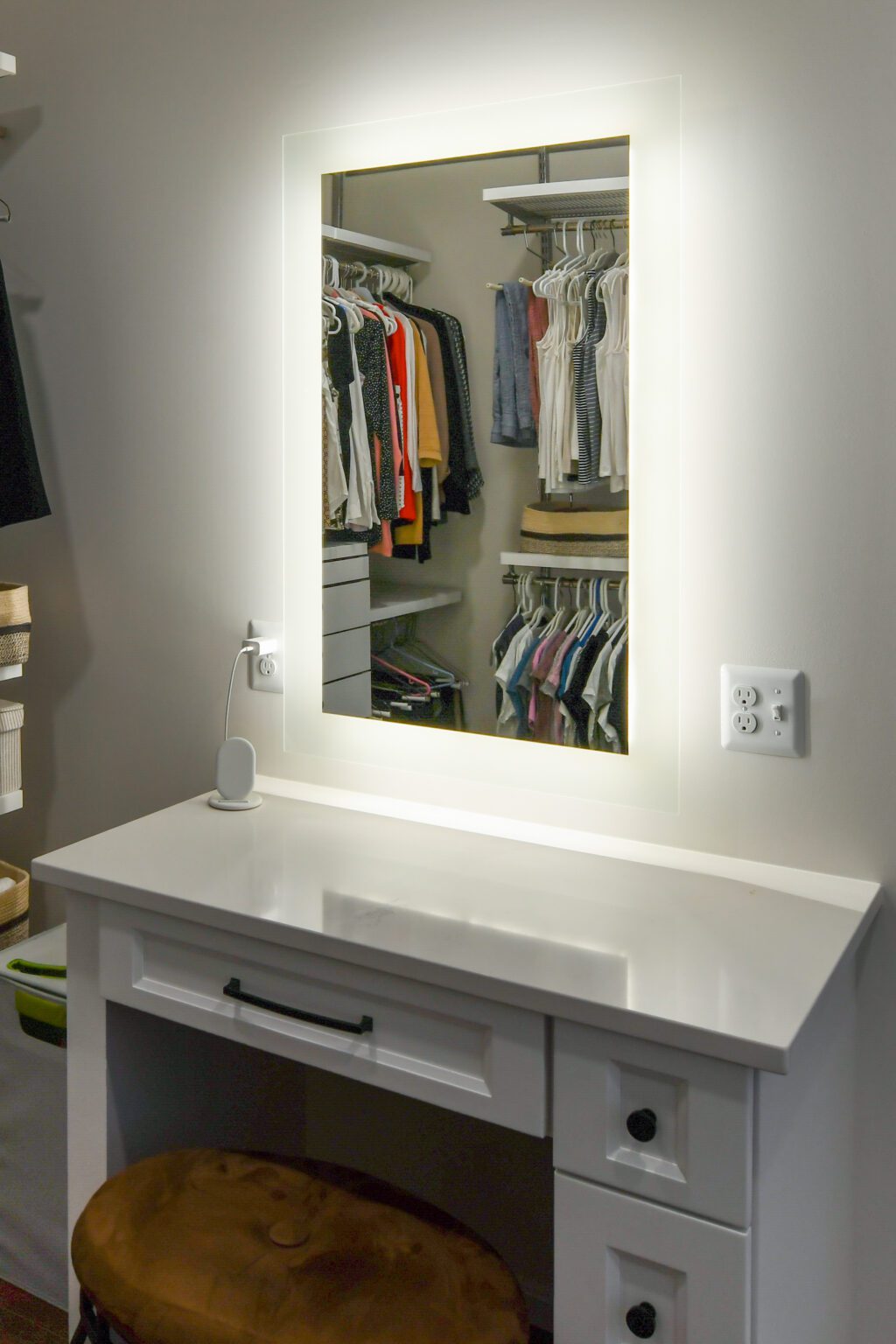 Vienna Primary En-suite and Laundry (15) A white vanity table with a brown stool sits below a large illuminated mirror, reflecting a walk-in closet with hanging clothes and shelves.