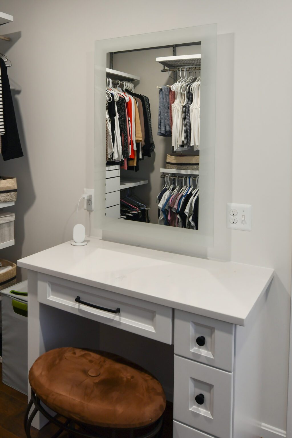 Vienna Primary En-suite and Laundry (16) A white vanity with a mirror and brown cushioned stool sits in front of a closet with hanging clothes visible in the reflection.