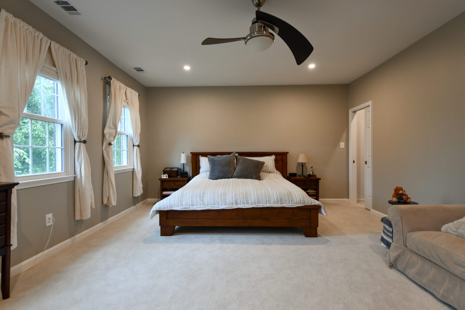 Vienna Primary En-suite and Laundry (22) A spacious bedroom with a large bed, neutral walls, two windows with curtains, ceiling fan, nightstands, and beige carpeted floor.