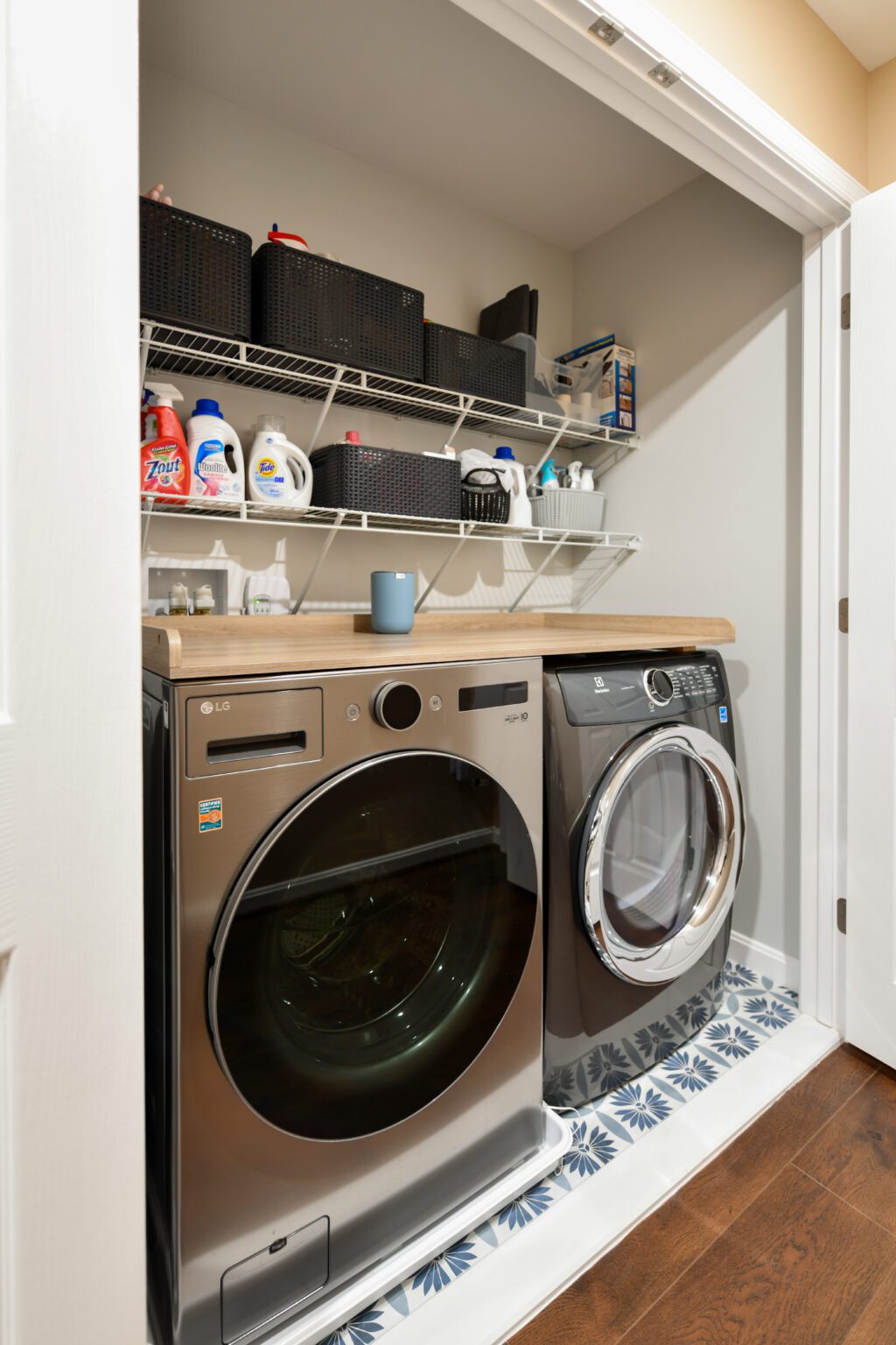 Vienna Primary En-suite and Laundry (24) Modern laundry closet with a front-loading washer and dryer, wooden countertop, shelving above holding baskets and laundry supplies, and patterned tile floor.