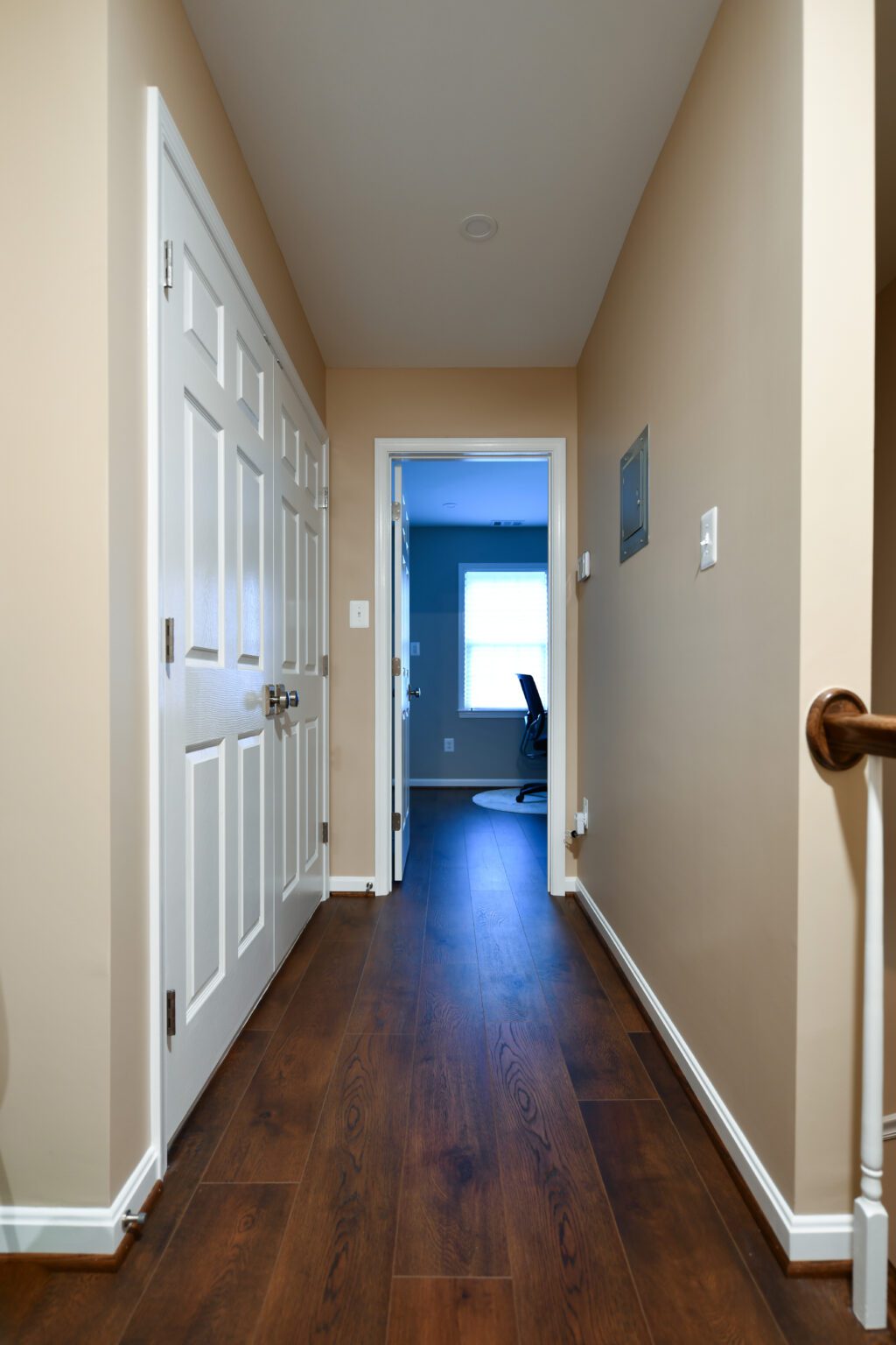 Vienna Primary En-suite and Laundry (28) A hallway with wooden flooring, beige walls, white doors on the left, and an office room with a chair visible at the end.