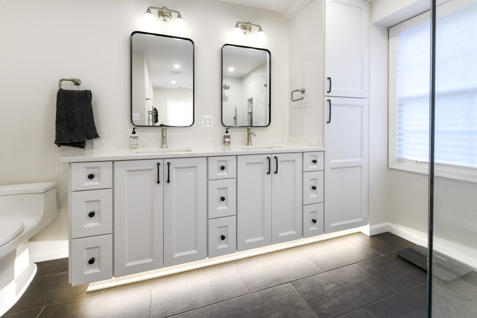 Vienna Primary En-suite and Laundry (3) Modern bathroom with double sink vanity, two mirrors, white cabinets, black fixtures, towel on wall, and under-cabinet lighting. Large window with blinds on the right side.