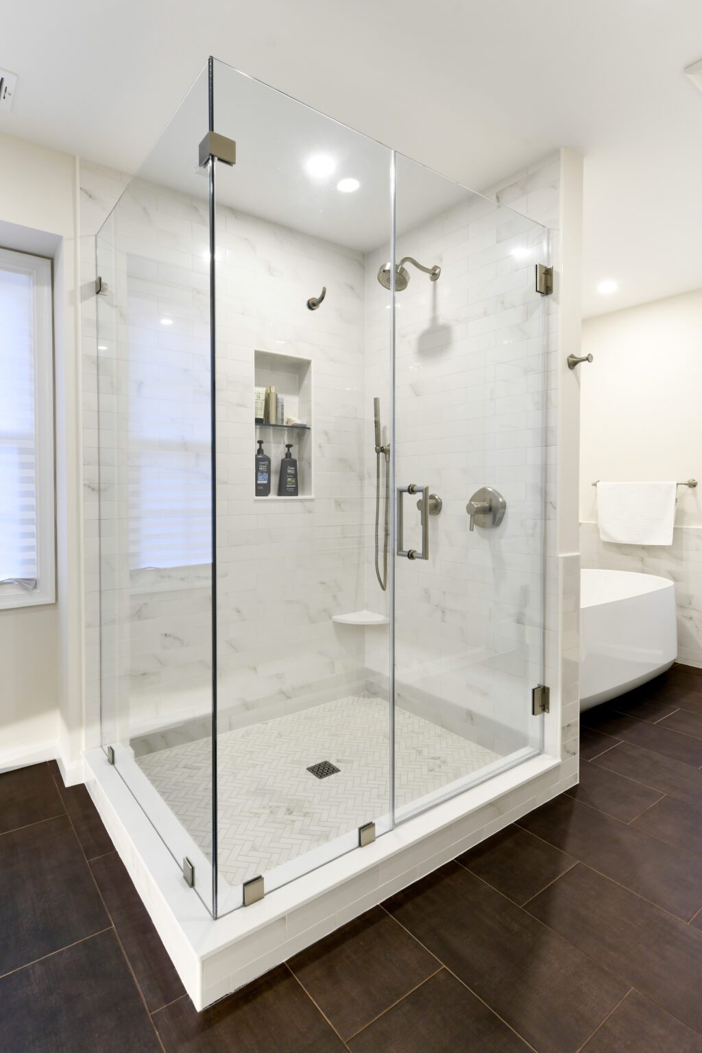 Vienna Primary En-suite and Laundry (4) A modern glass-enclosed shower with white tile walls, a built-in shelf holding bottles, a bench, and a rainfall showerhead, set on a dark tile floor.