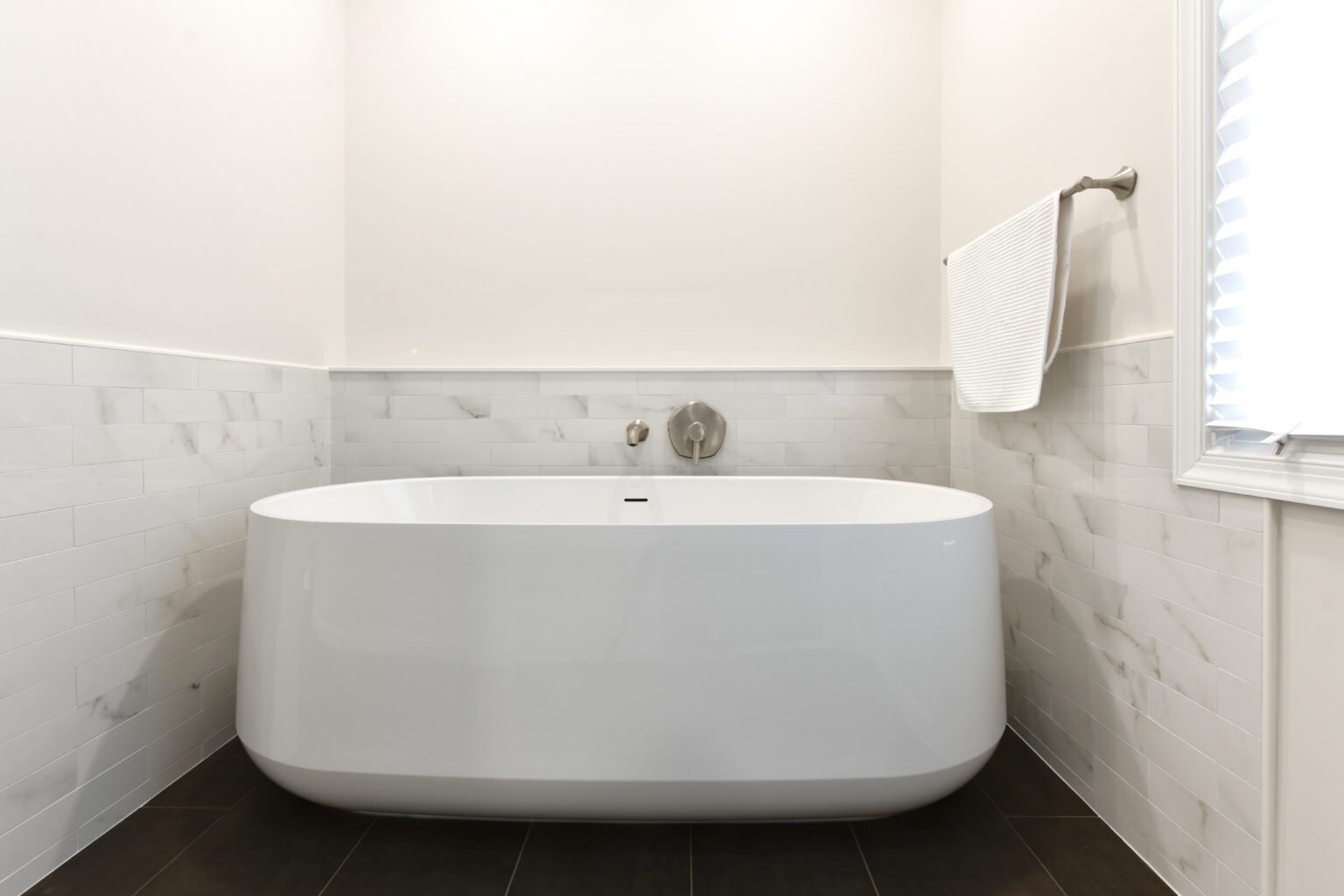 Vienna Primary En-suite and Laundry (6) Modern white bathtub in a minimalist bathroom with light-colored tiled walls, dark floor tiles, a wall-mounted faucet, and a towel hanging on a rod.
